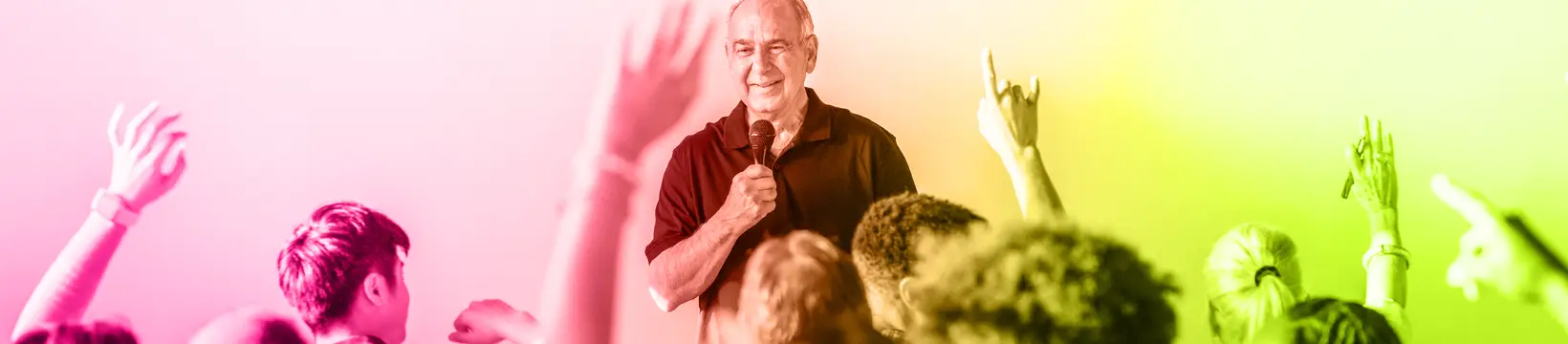 Mann mit Mikrofon vor einer Gruppe von Personen von denen einige sich melden. Bild ist mit einem Farbverlauf aus consus healthcare akademie Farben eingefärbt.